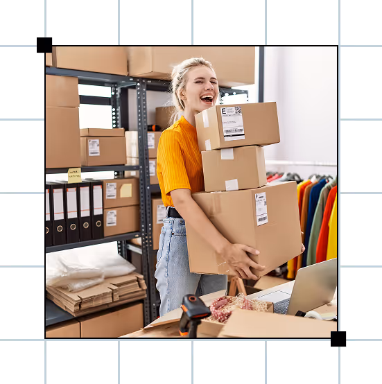 Person smiling while carrying multiple shipping boxes in a small warehouse workspace.