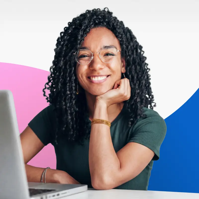 Smiling woman with curly hair and glasses sitting at a desk with a laptop, resting her chin on her hand.