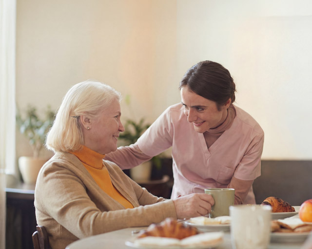 A caregiver smiling and holding a cup while interacting warmly with an elderly woman seated at a table with breakfast items.