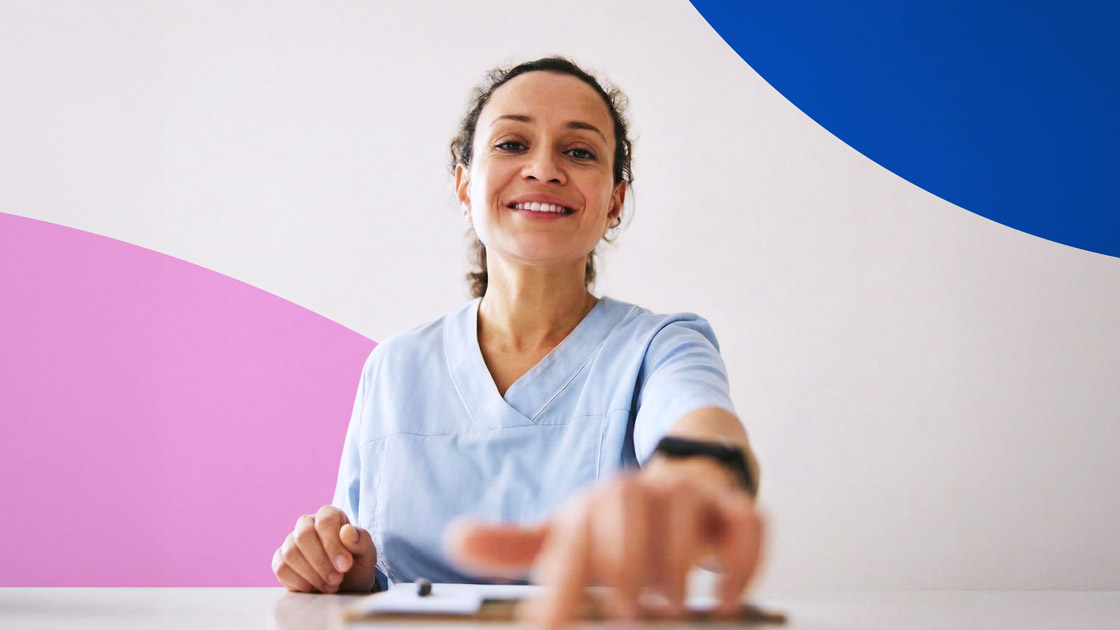Smiling healthcare worker in light blue scrubs reaching forward with hand toward the camera against a colorful abstract background.
