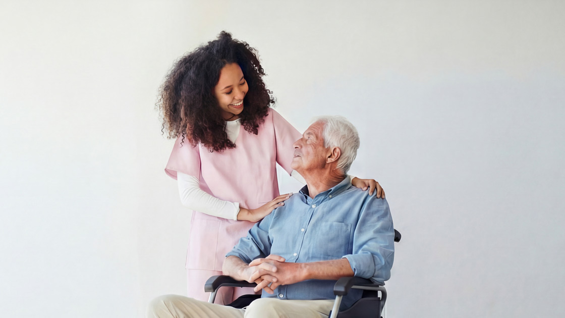 Caregiver in pink uniform smiling and comforting an elderly man sitting in a wheelchair against a plain background.