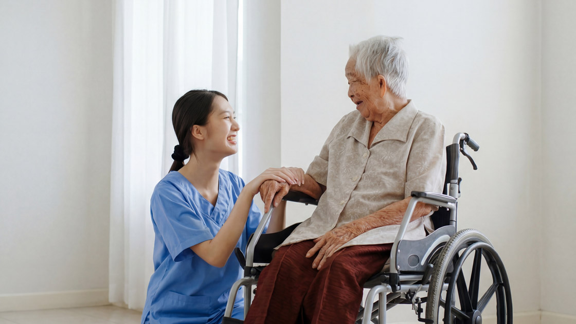 Smiling caregiver in blue scrubs holding hands and interacting warmly with elderly woman in a wheelchair.