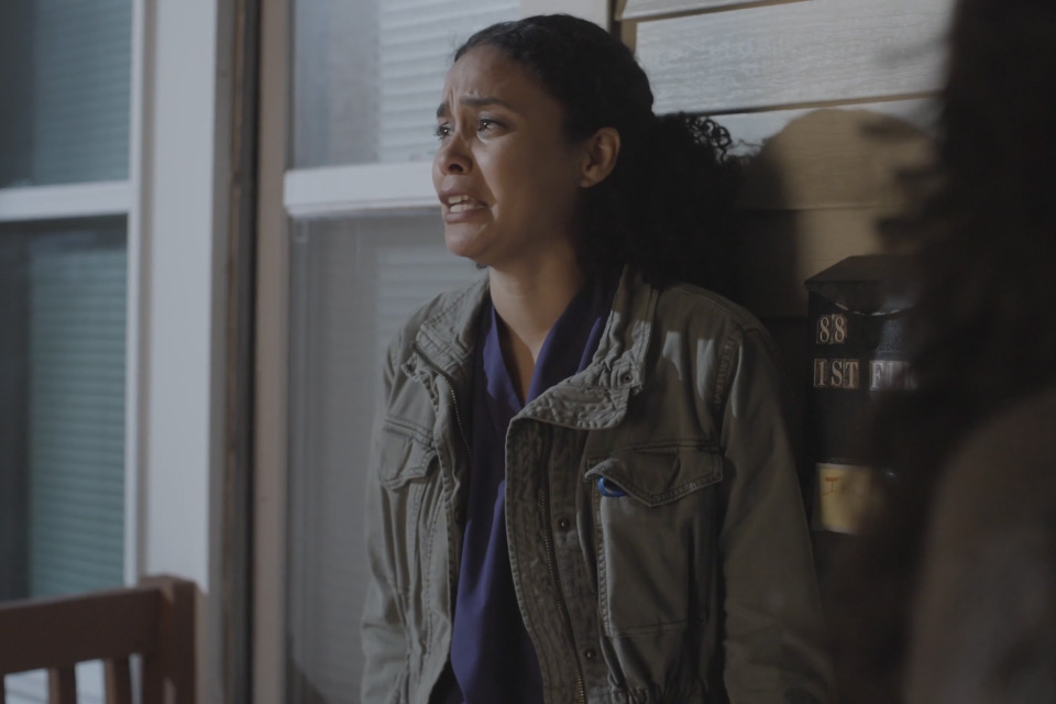 A woman with curly hair and a green jacket looks distressed, standing against a wall near a window and a mailbox.