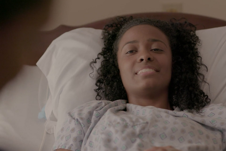 Smiling woman with curly hair lying in a hospital bed wearing a patterned hospital gown.