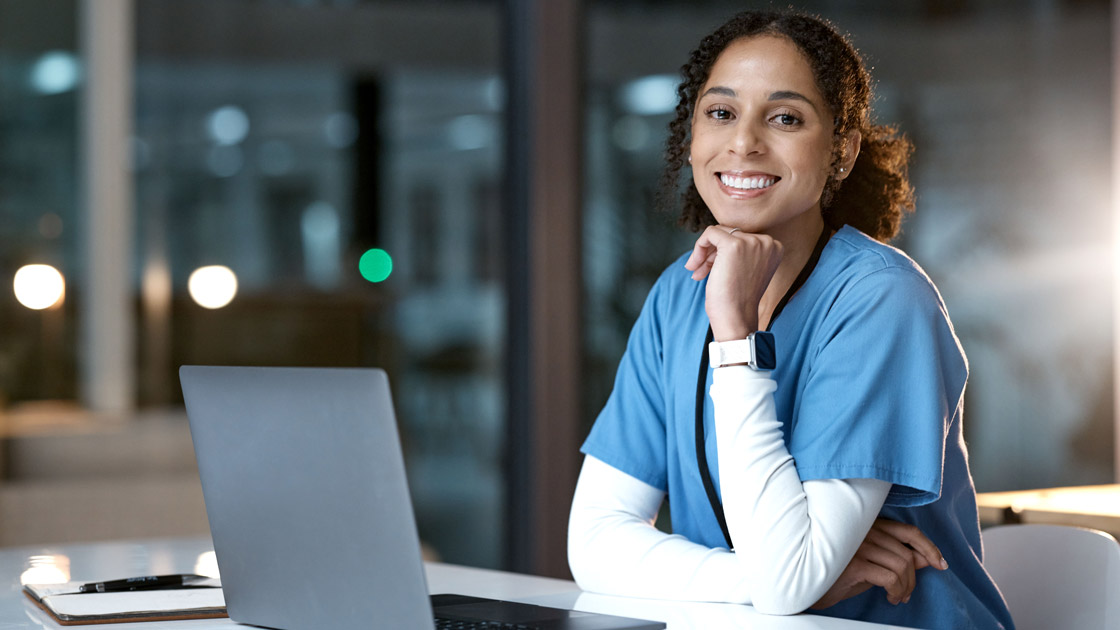 Smiling female nurse in blue scrubs sitting at a desk with a laptop and clipboard indoors.