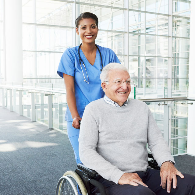 Smiling nurse in blue scrubs pushing an elderly man in a wheelchair through a modern building corridor.