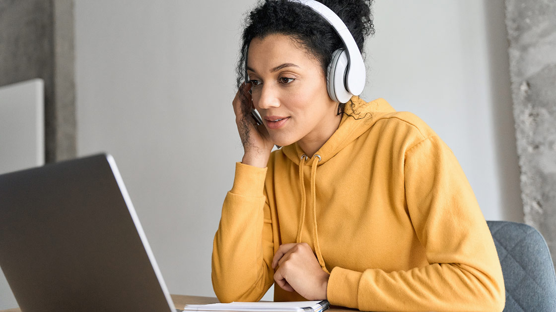 Young woman in a yellow hoodie wearing white headphones and looking attentively at a laptop screen.