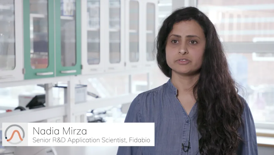A woman with long, dark hair stands in a lab setting, wearing a blue blouse, with laboratory equipment and cabinets in the background.