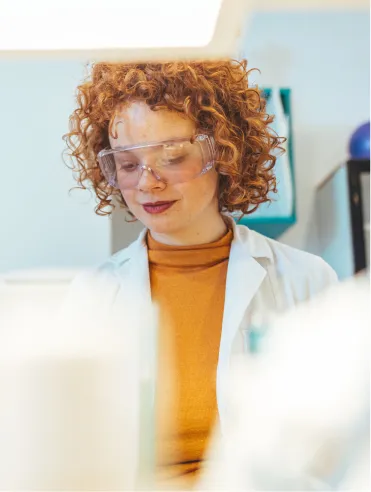 Young woman with curly red hair wearing safety goggles, a white lab coat, and an orange turtleneck in a laboratory setting.