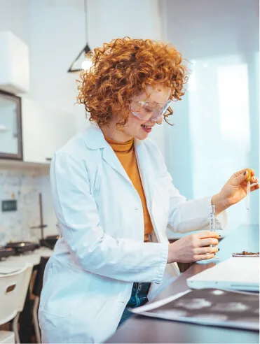 Woman with curly red hair wearing safety goggles and a white lab coat conducts an experiment with a liquid dropper in a bright laboratory.