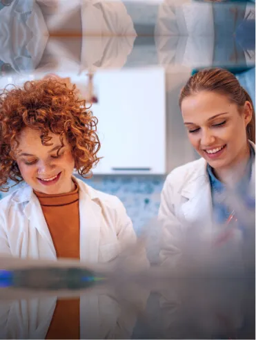 Two female scientists in white lab coats smiling and working together in a laboratory.