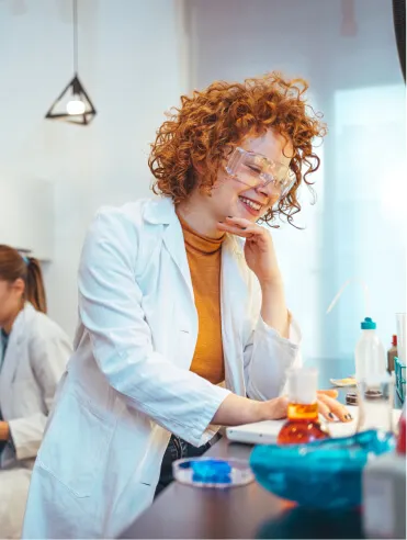Smiling female scientist with curly red hair and safety glasses working at a lab bench.
