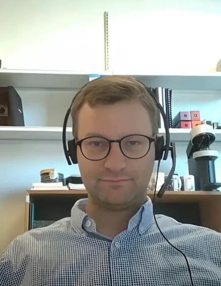 Man wearing glasses and a headset seated in front of shelves with office supplies.