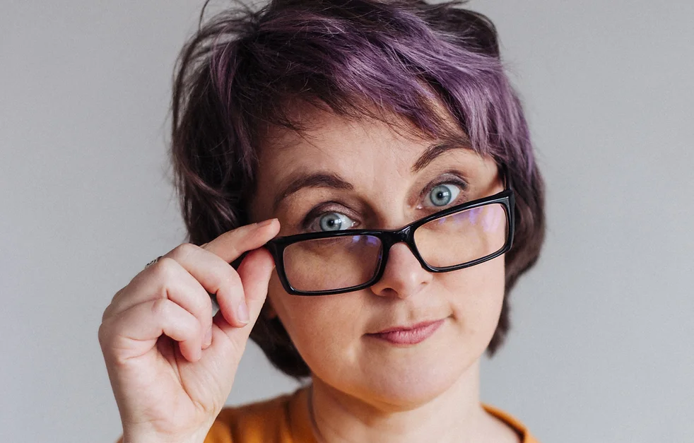 Портрет Оксаны Шапкариной. Её волосы окрашены в фиолетовый, глаза серые. Она внимательно смотрит на зрителя, приспустив рукой очки в тёмной прямоугольной оправе, на губах лёгкая улыбка