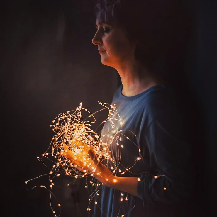 Женщина, стоящая в темноте, держит в руках светящуюся гирлянду с маленькими жёлтыми огоньками