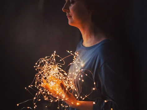 Женщина, стоящая в темноте, держит в руках светящуюся гирлянду с маленькими жёлтыми огоньками