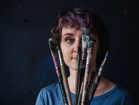 Оксана Шапкарина держит веером кисти для рисования и смотрит на зрителя через них