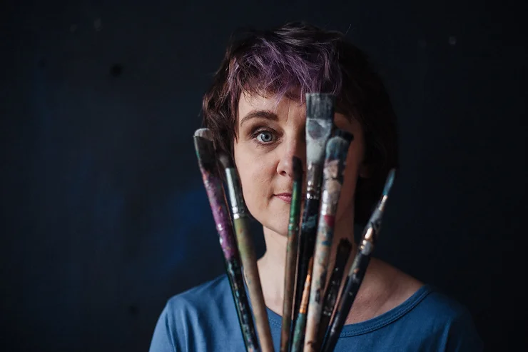 Оксана Шапкарина держит веером кисти для рисования и смотрит на зрителя через них