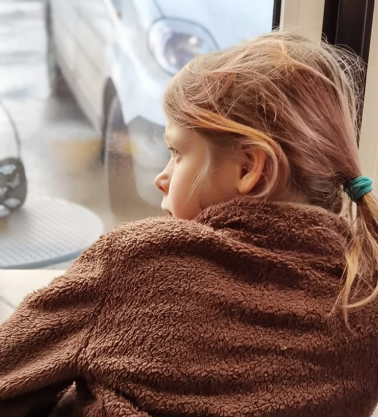 Девочка во флисовой коричневой куртке смотрит в окно, стоя спиной к зрителю