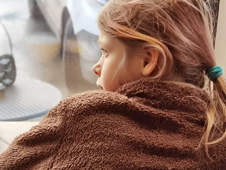 Девочка во флисовой коричневой куртке смотрит в окно, стоя спиной к зрителю