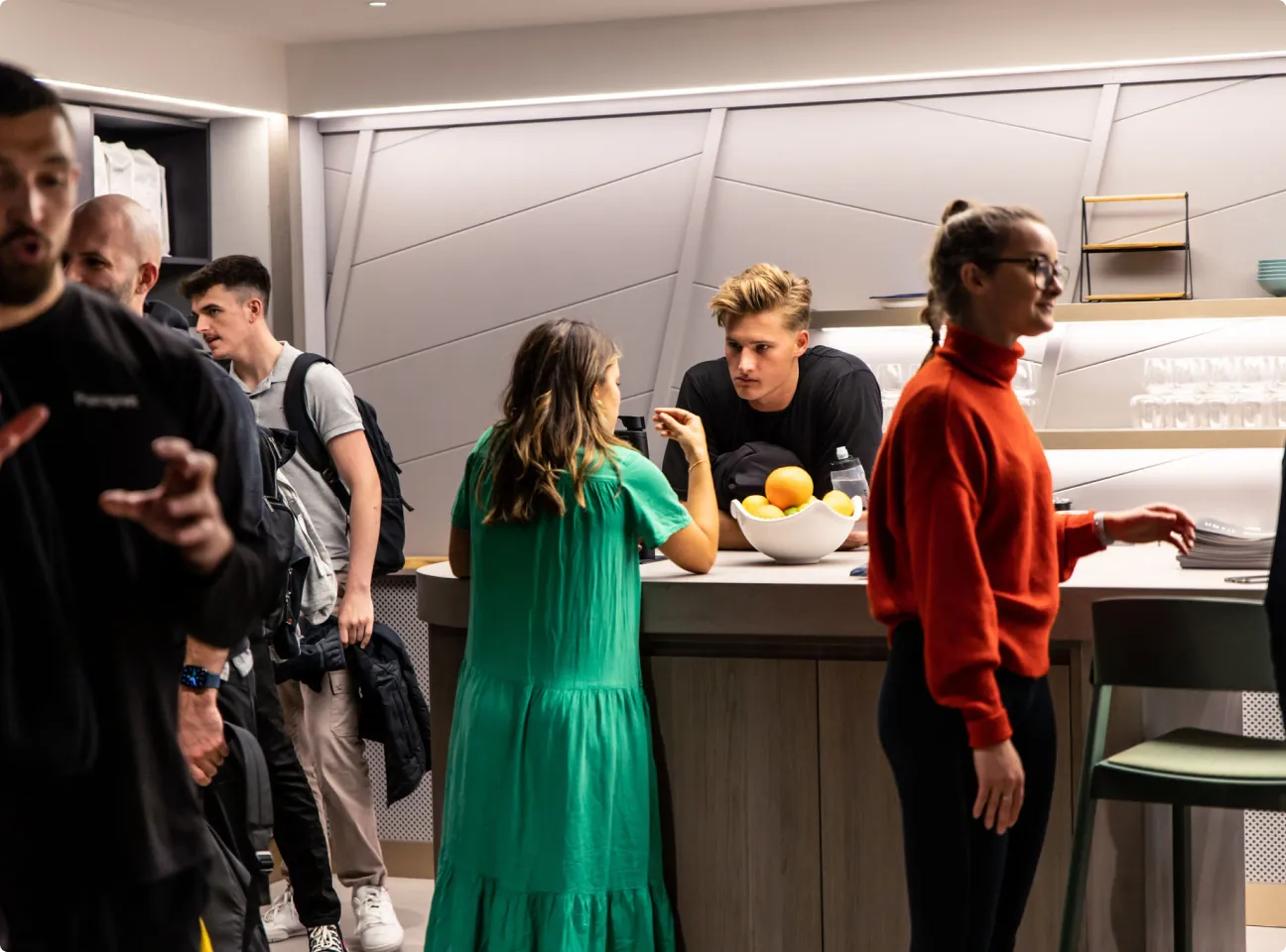 Group of young adults socializing near a modern kitchen counter with a bowl of fruit.