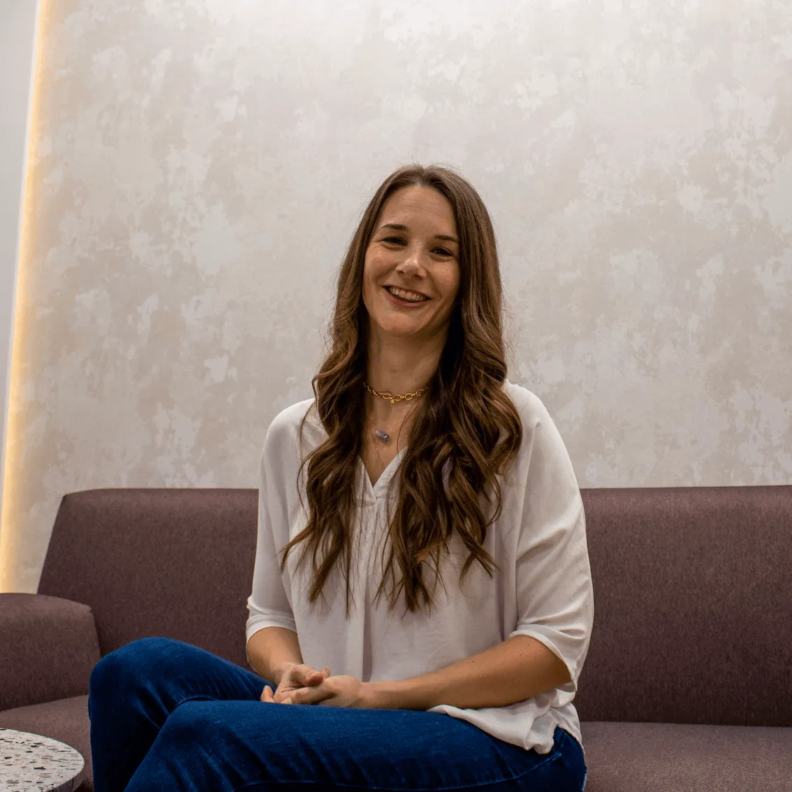 Smiling woman with long wavy hair wearing a white blouse and blue jeans sitting on a brown sofa.