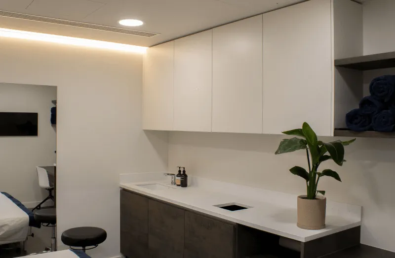 Modern, clean room with a white countertop, brown lower cabinets, white upper cabinets, potted plant, hand soap, and rolled navy towels on shelves.
