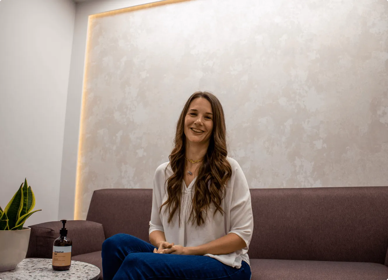 Smiling woman with long wavy brown hair sitting cross-legged on a purple couch in a modern room with a plant and hand sanitizer on a round table.