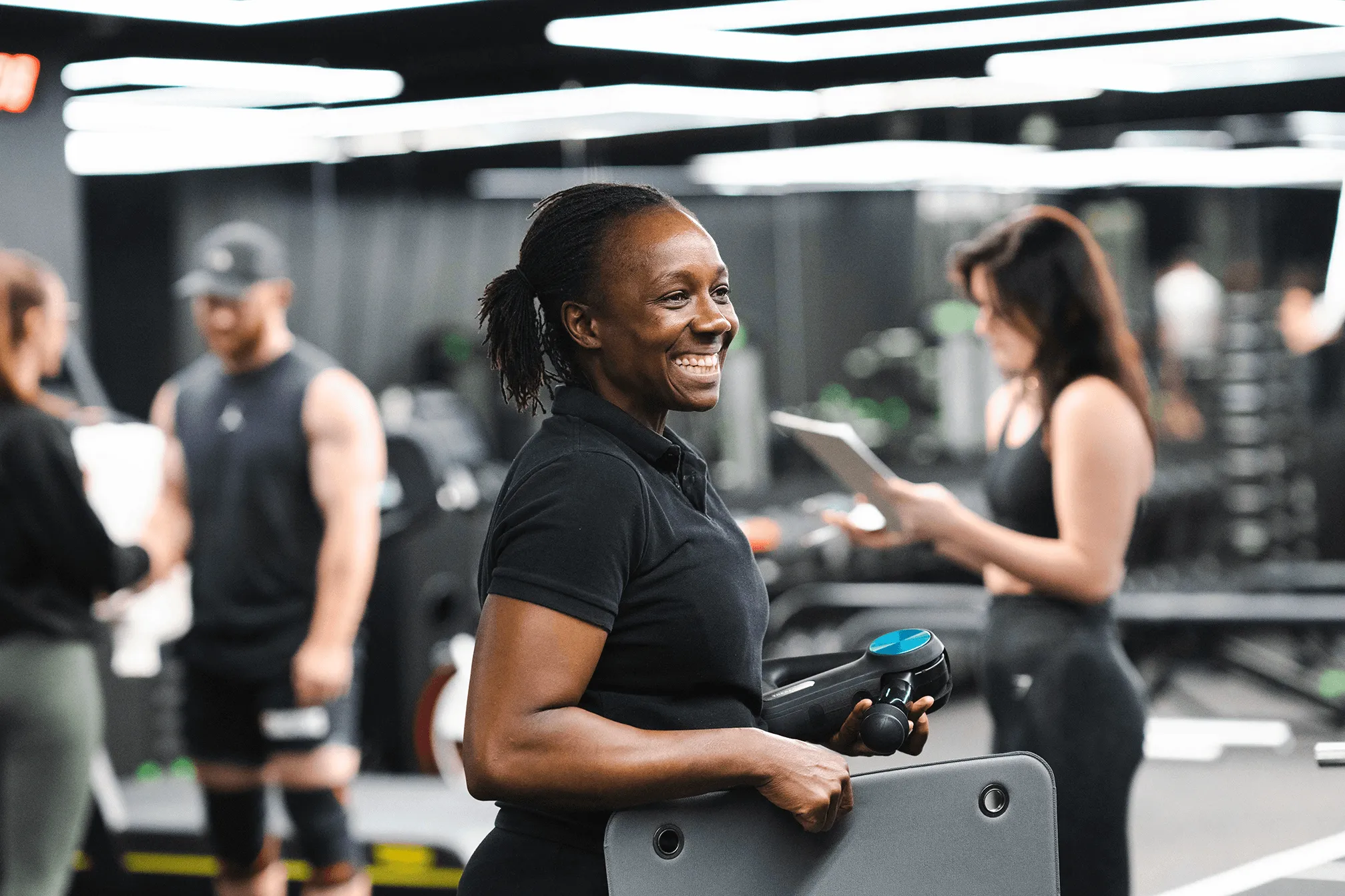 Smiling woman in black shirt holding a handheld massage gun and a workout mat in a gym with blurred people in the background.