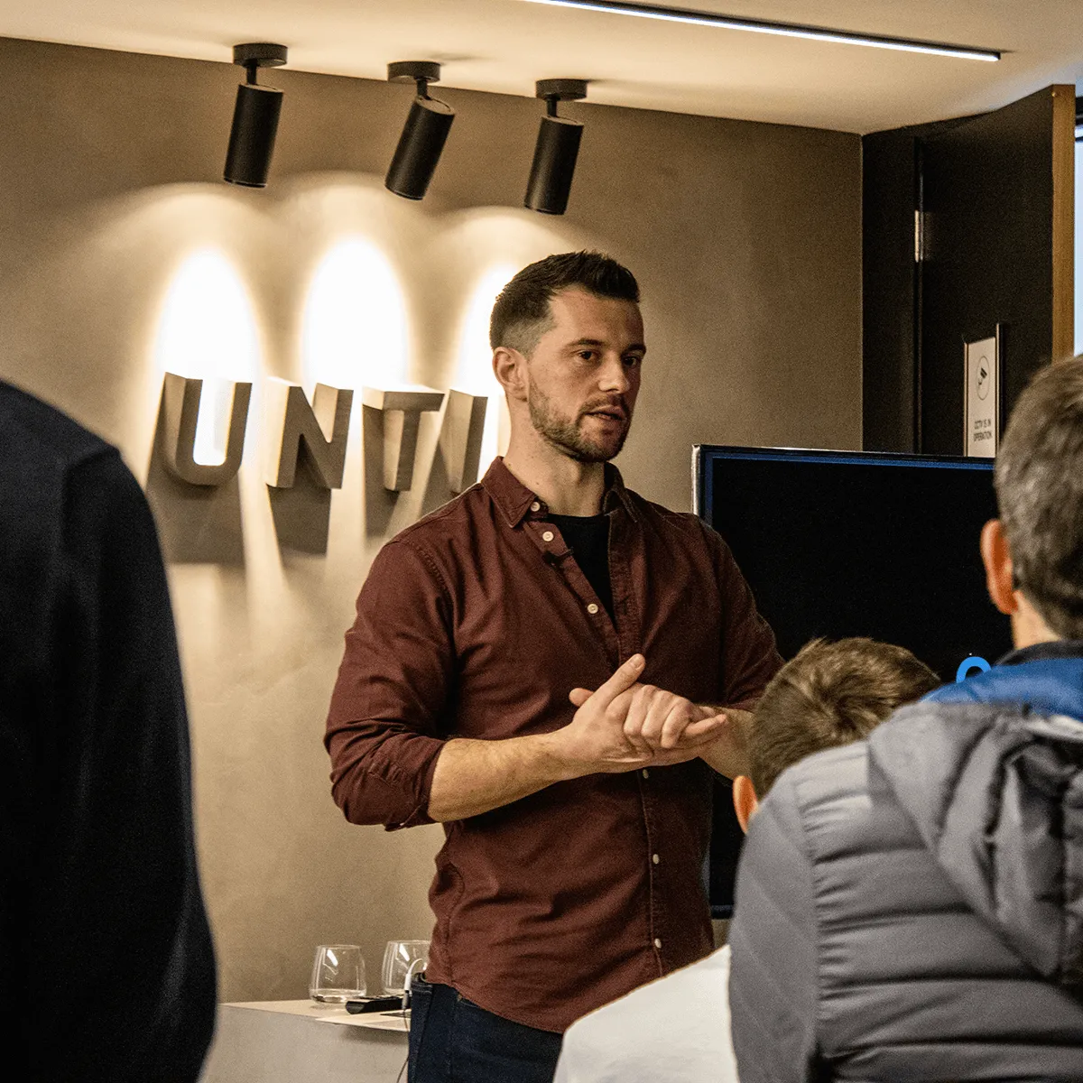 Man in a brown shirt speaking to an audience in a modern room with spotlighted wall letters.