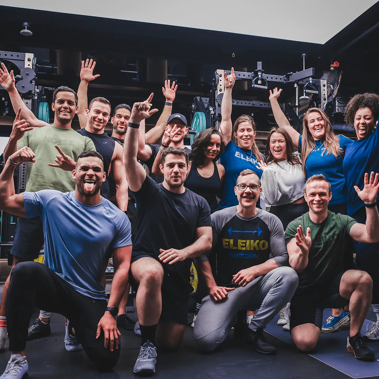 Group of smiling men and women posing energetically in a gym setting with weightlifting equipment in the background.