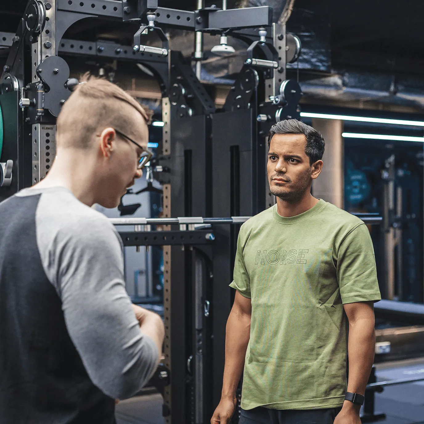 Two men talking in a gym with weightlifting equipment in the background, one wearing glasses and a gray shirt, the other wearing a green t-shirt.