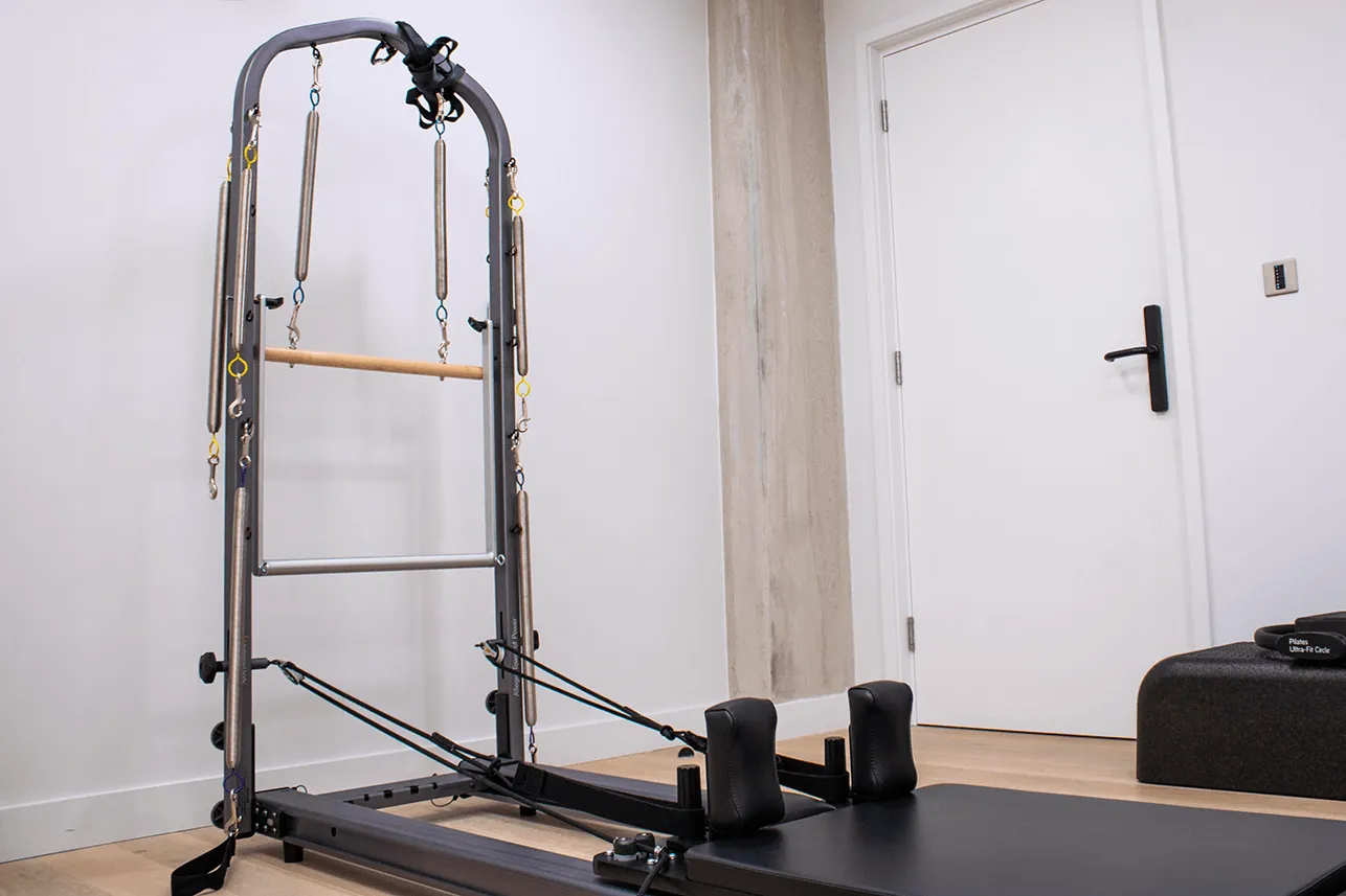 Pilates reformer machine with springs and foot straps in a bright exercise room.