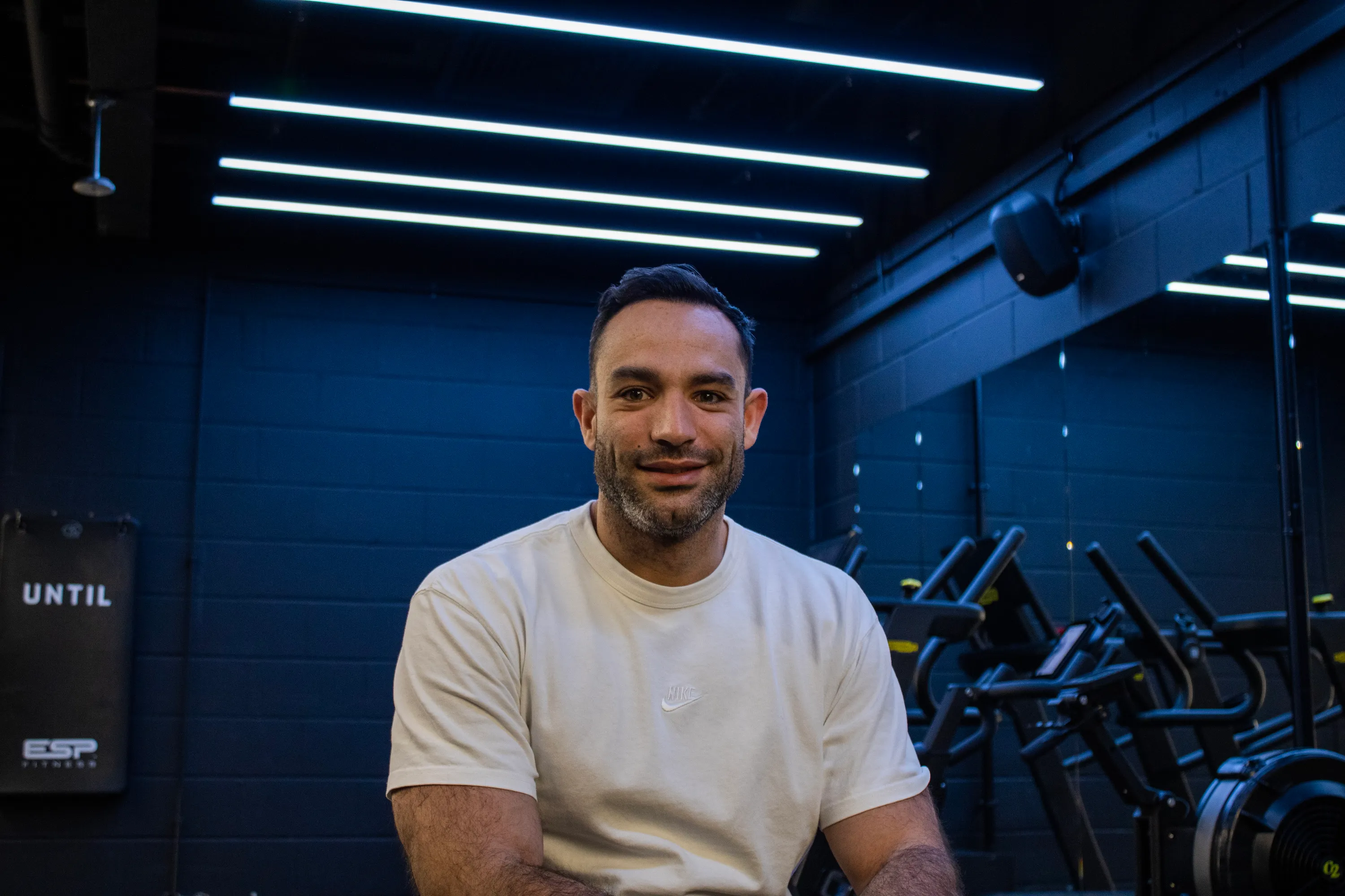 Man in a white Nike t-shirt sitting in a gym with exercise equipment and blue walls in the background.