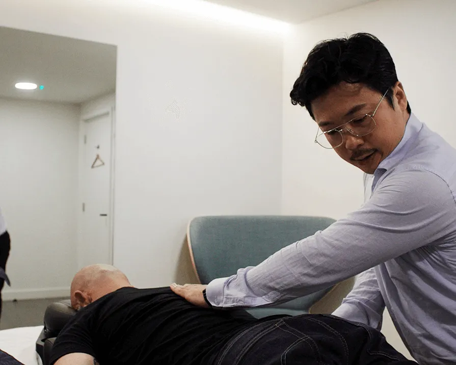 Male therapist in glasses and light purple shirt performing a back massage on a bald man lying face down on a treatment table in a modern room.