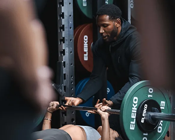 Male trainer spotting a woman doing bench press with a barbell loaded with Eleiko weight plates in a gym.