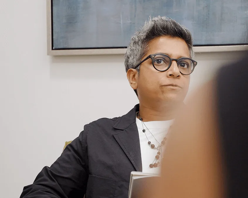 Person with short gray hair and glasses wearing a black jacket and white shirt, sitting and looking thoughtfully.