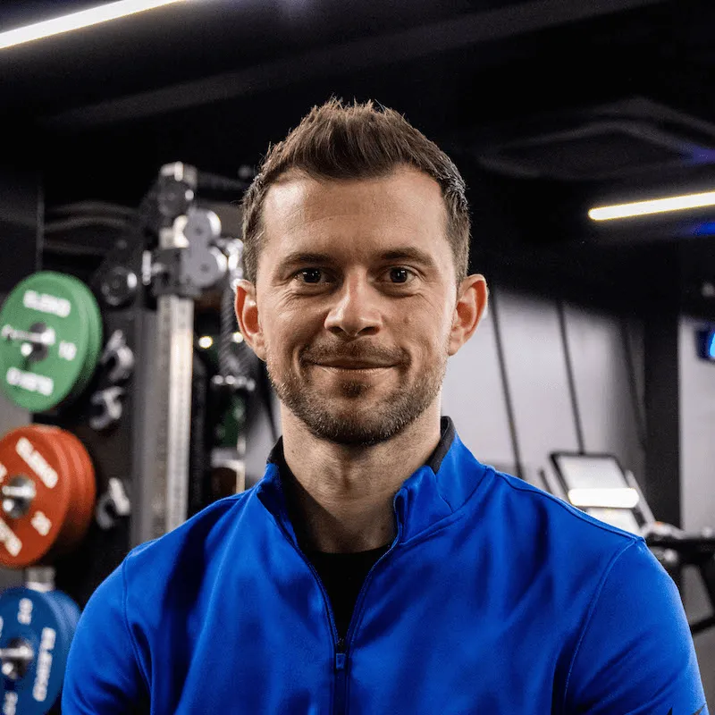 Smiling man with short brown hair wearing a blue zip-up athletic jacket in a gym setting with weight plates in the background.