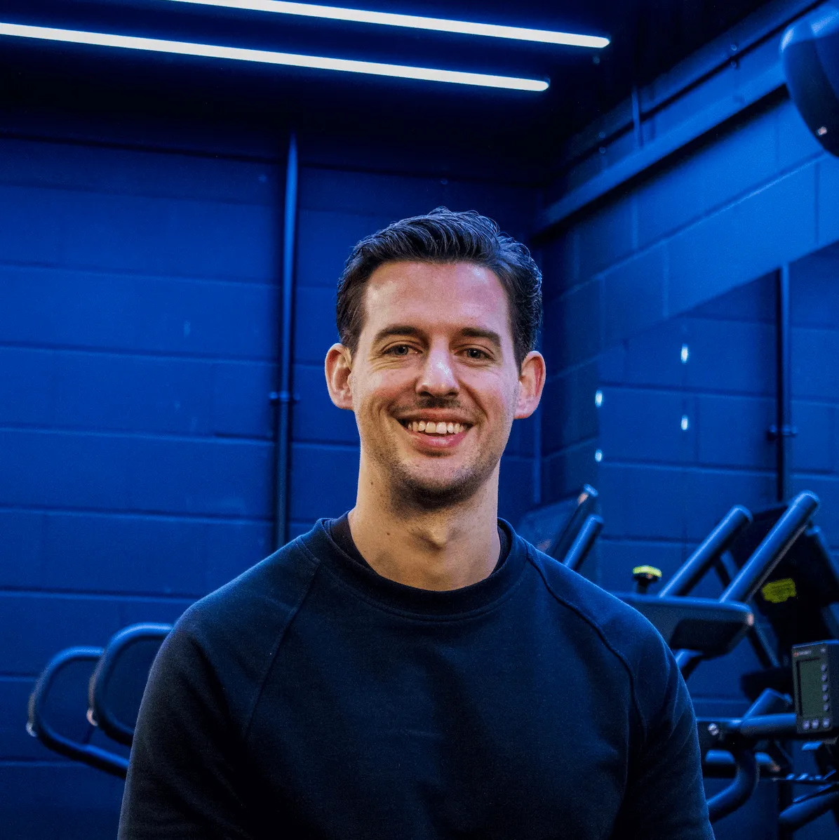 Smiling man with short dark hair wearing a black sweatshirt in a gym setting with exercise equipment and blue walls.