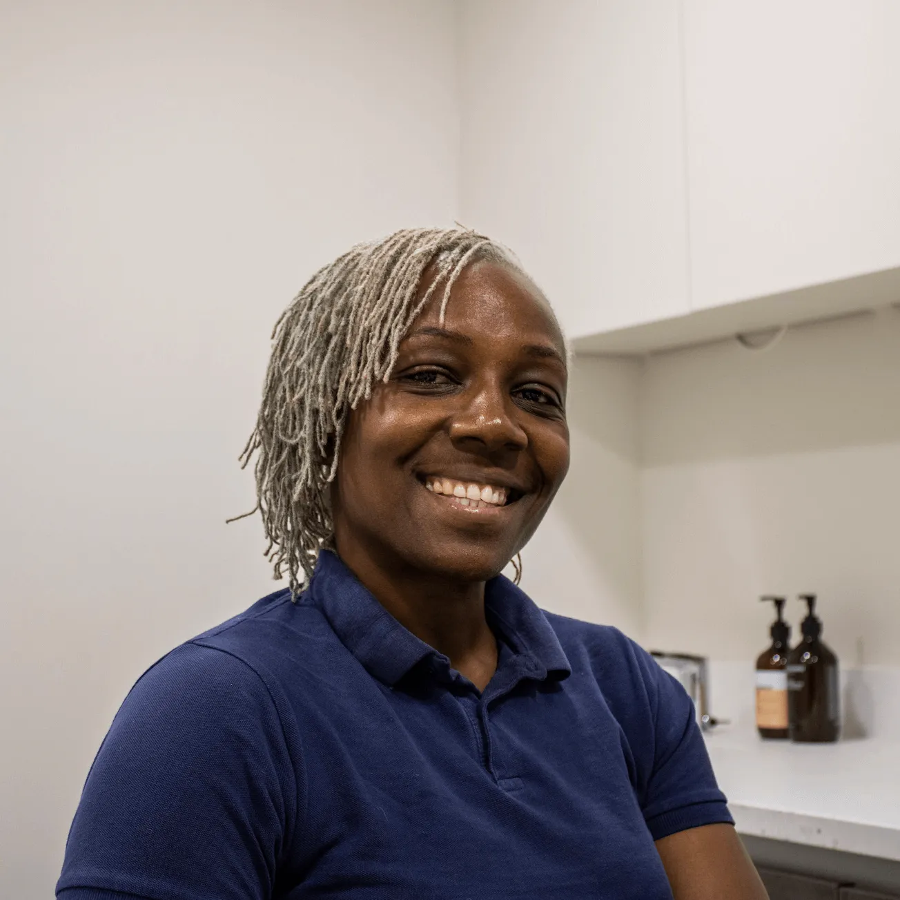 Smiling woman with short gray dreadlocks wearing a navy blue polo shirt in a room with white walls and countertop.
