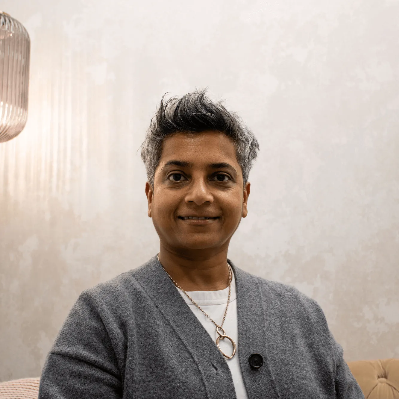 Smiling person with short salt-and-pepper hair wearing a grey cardigan, white shirt, and a pendant necklace, sitting indoors against a textured light wall.