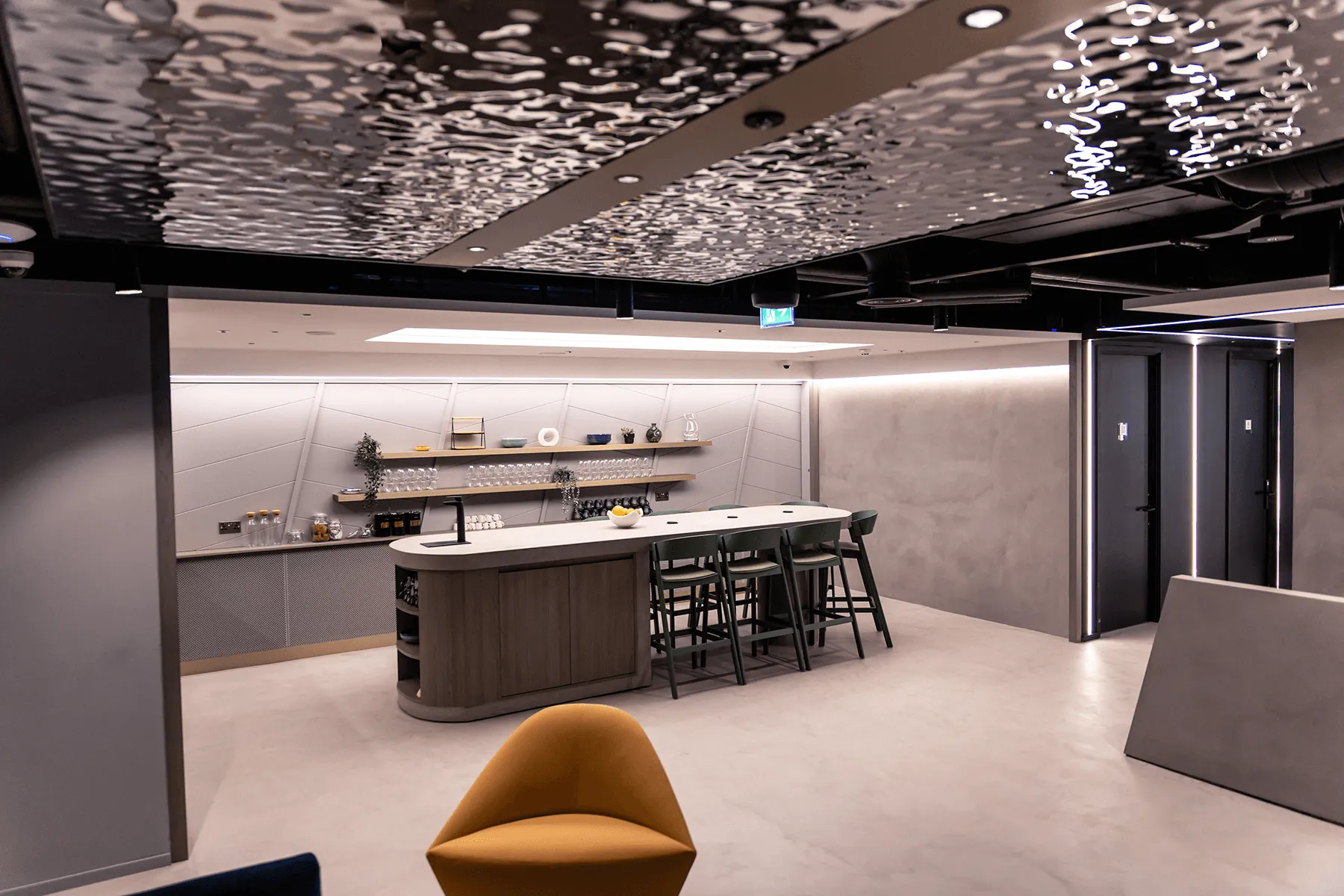 Modern office lounge with a central island bar, high stools, open shelves with glasses, and textured ceiling lighting.