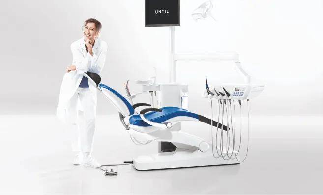Smiling female dentist in white coat standing beside modern dental chair with equipment in a bright, clean dental office.