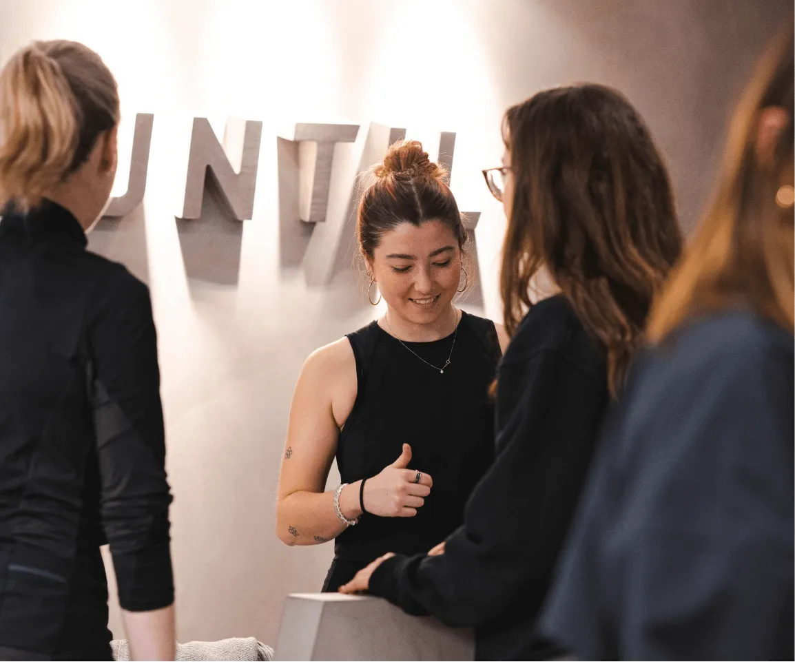 Smiling woman in a black sleeveless top interacting with two other women indoors, with partial large letters spelling 'DENTAL' on the wall behind them.