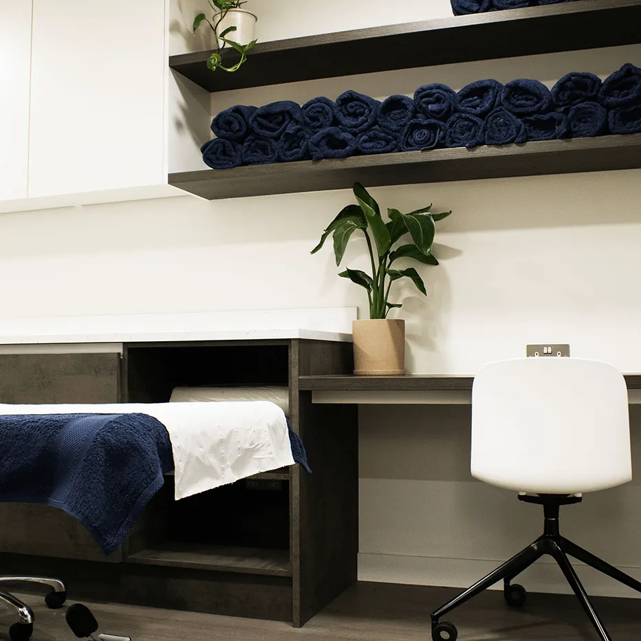Massage or treatment room with a table covered in white and navy towels, a white rolling chair, shelves with rolled navy towels, and a potted plant on a wooden desk.