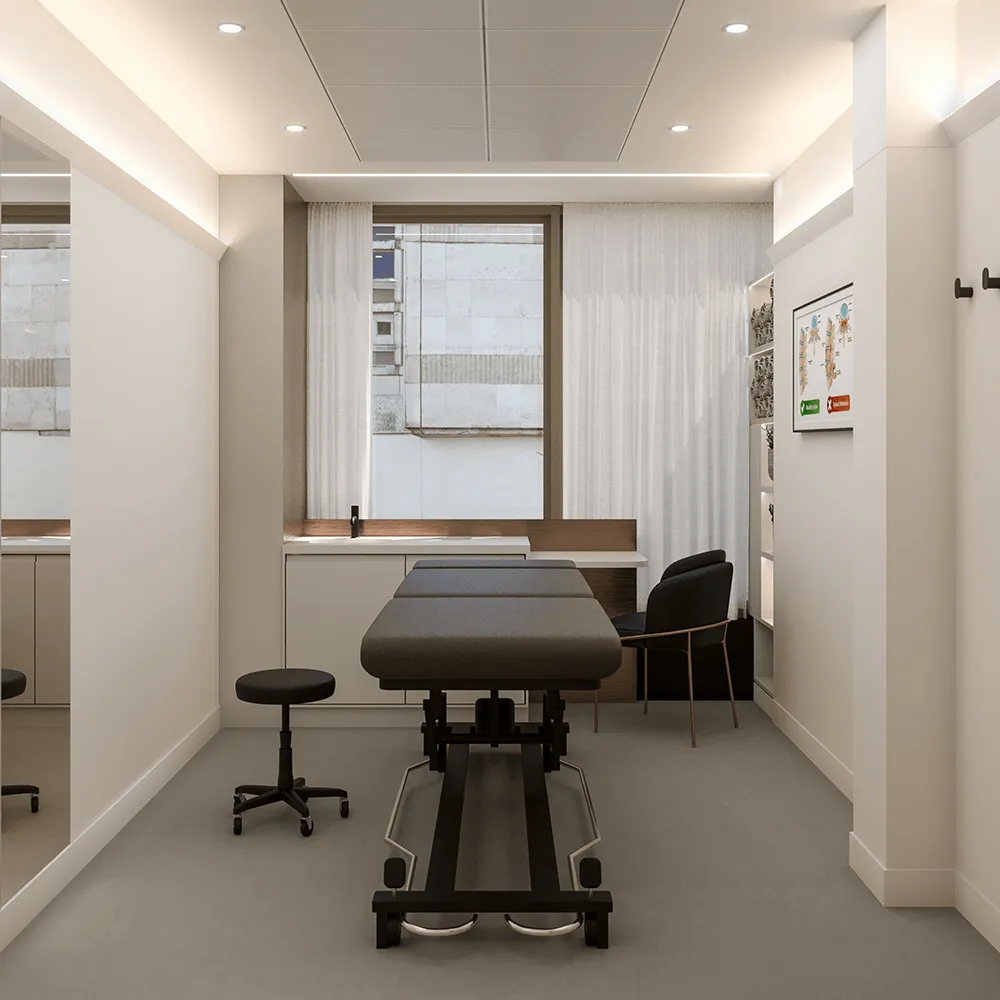 Modern therapy or treatment room with adjustable black treatment table, black rolling stool, armchair, sink, and anatomical chart on the wall.