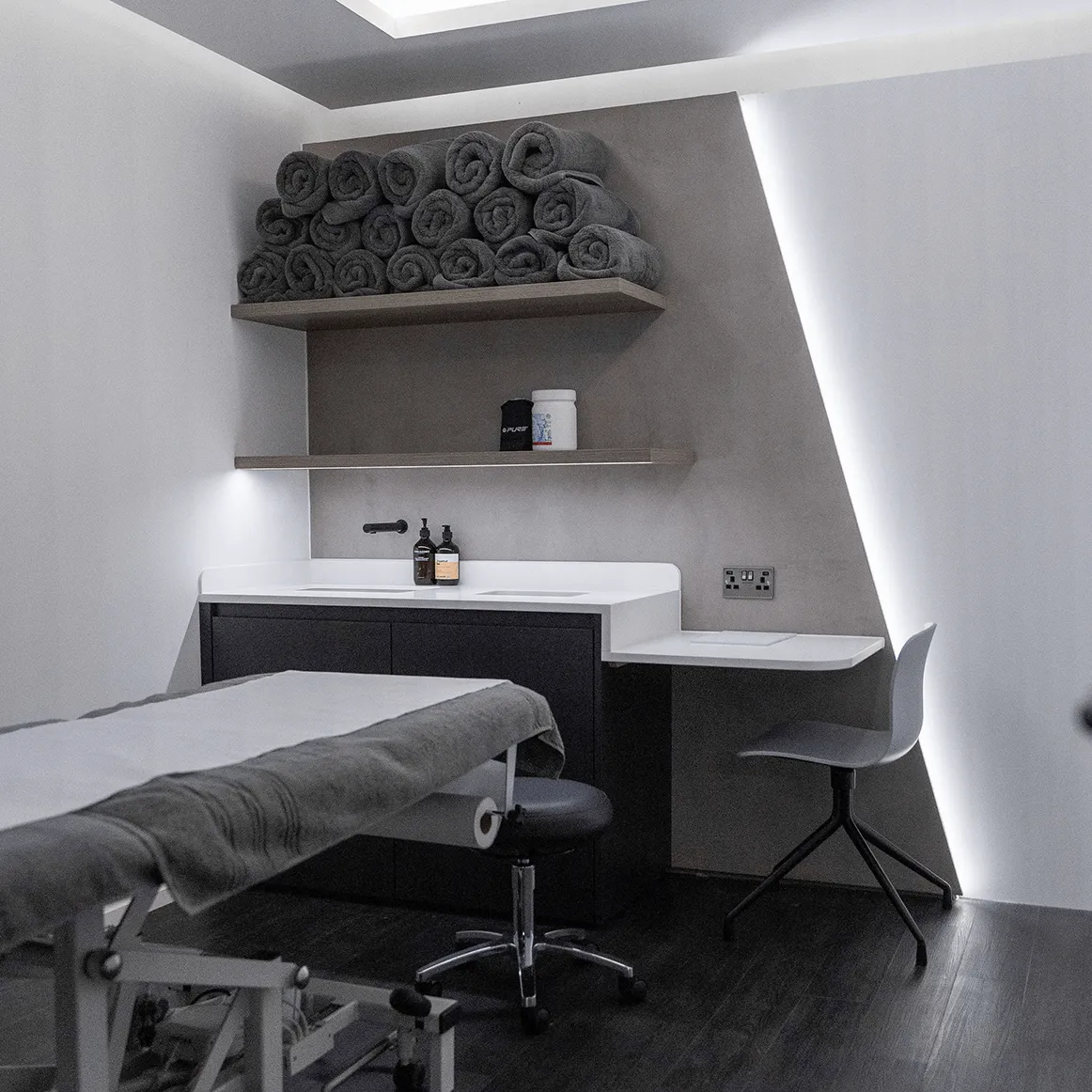 Modern spa treatment room with a massage table, rolled towels on a shelf, and a small desk with a chair.