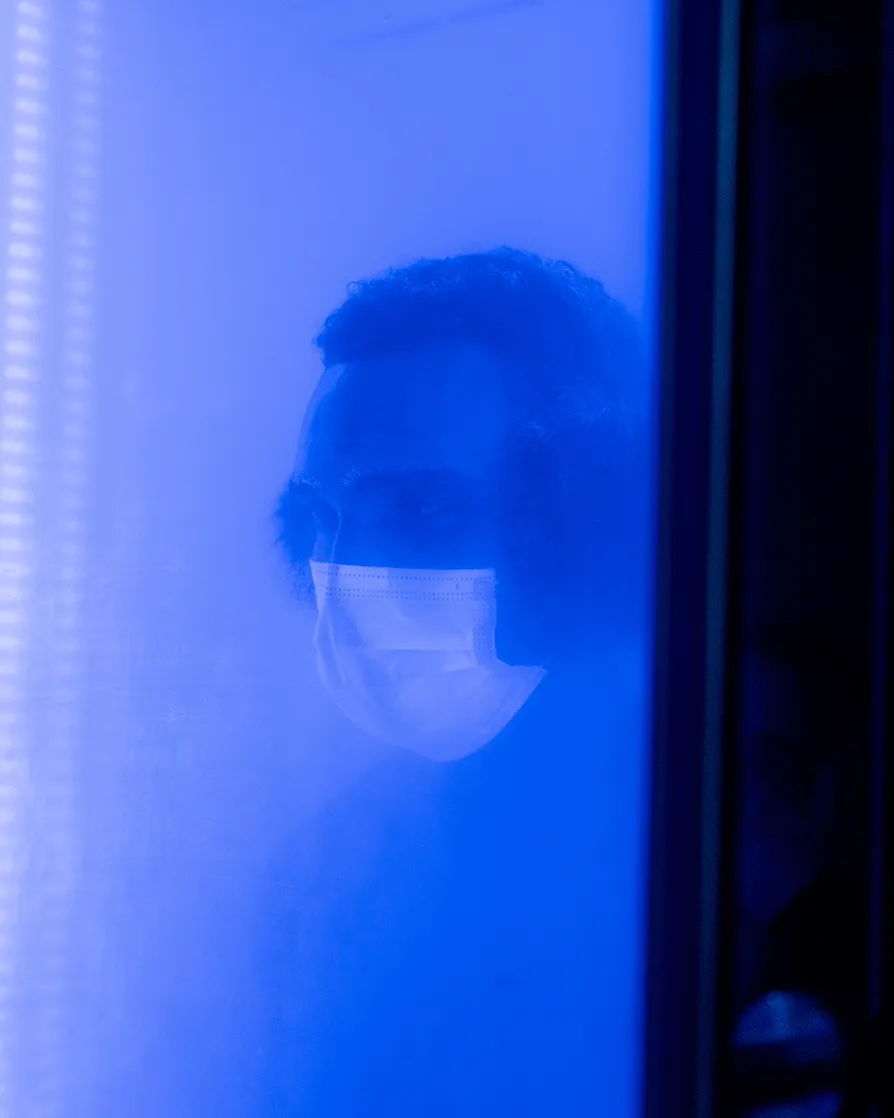 Person wearing earmuffs and a face mask, seen through a frosted blue-tinted glass.