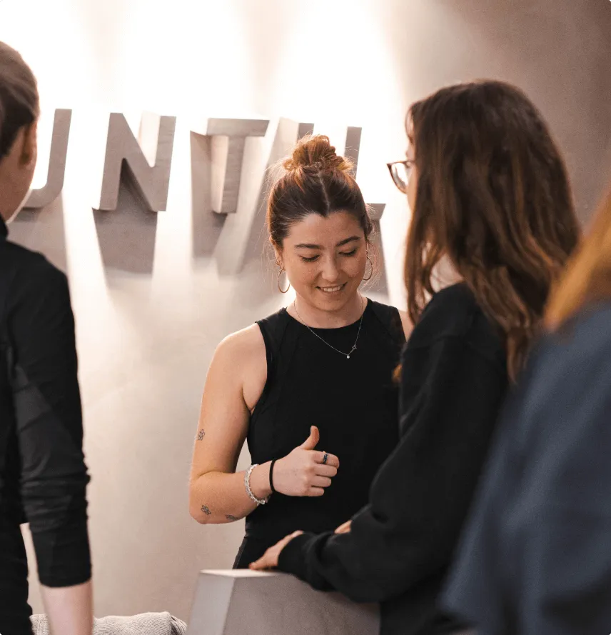 Woman with a black sleeveless top giving a thumbs-up while interacting with two other people indoors near a wall with large gray letters.