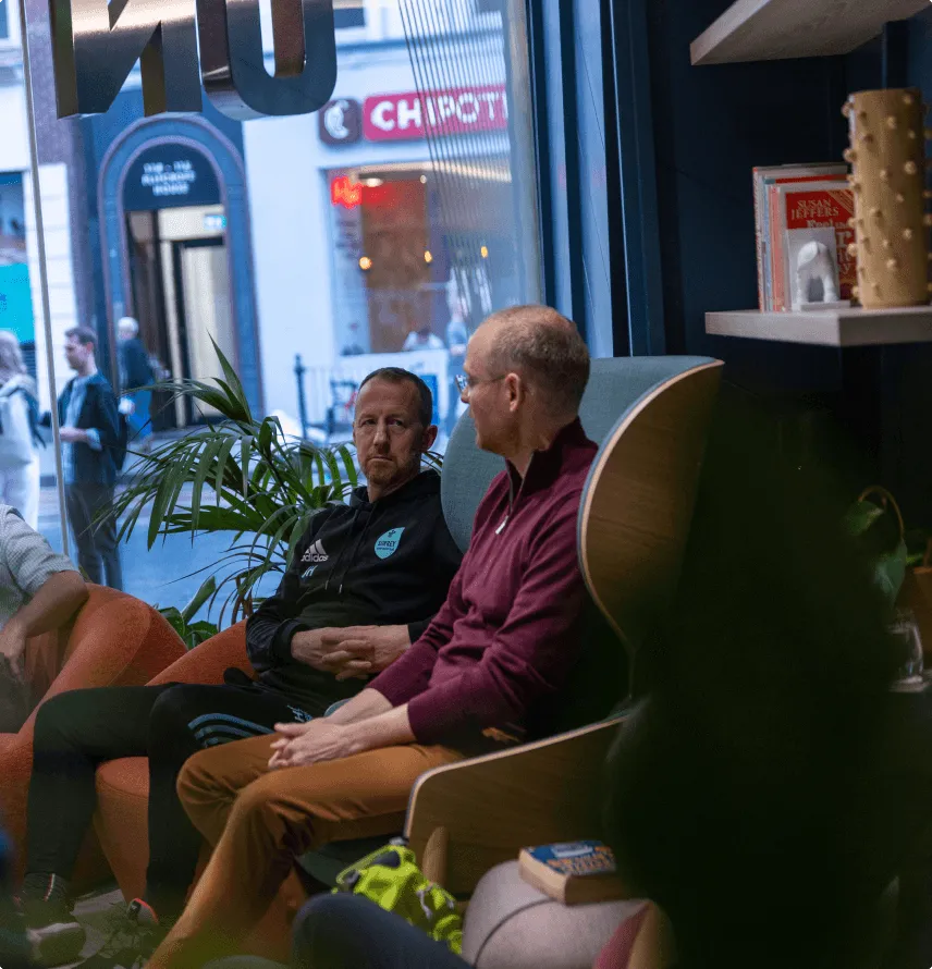 Two men sitting and conversing in a casual indoor setting near a large window with a view of people outside and a Chipotle sign.